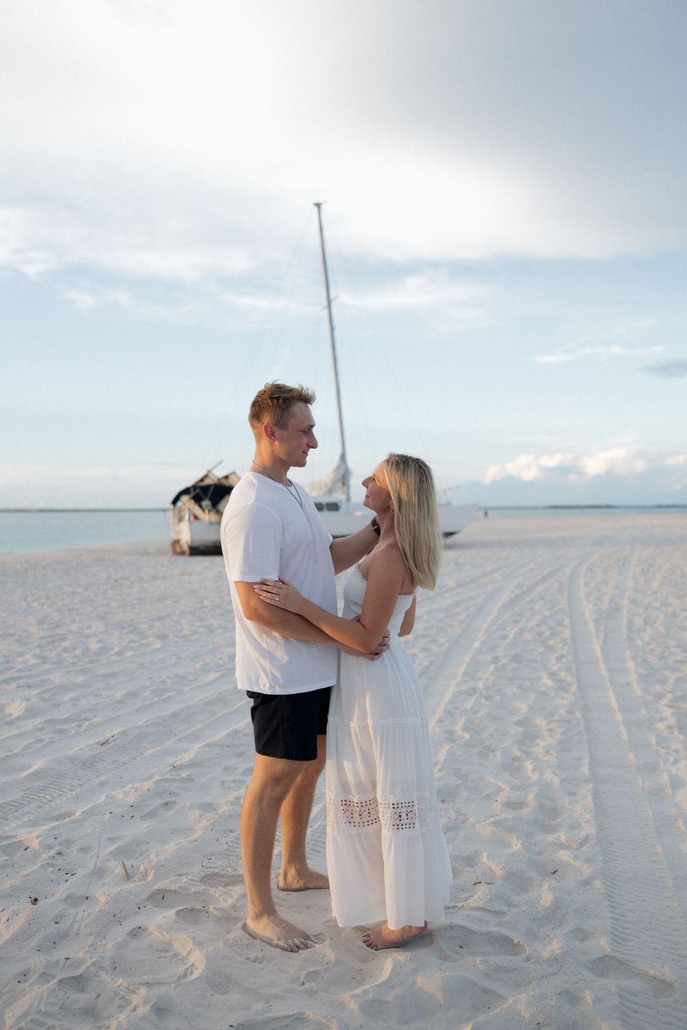 A couple embraces on a sandy beach at sunset with a sailboat in the background.