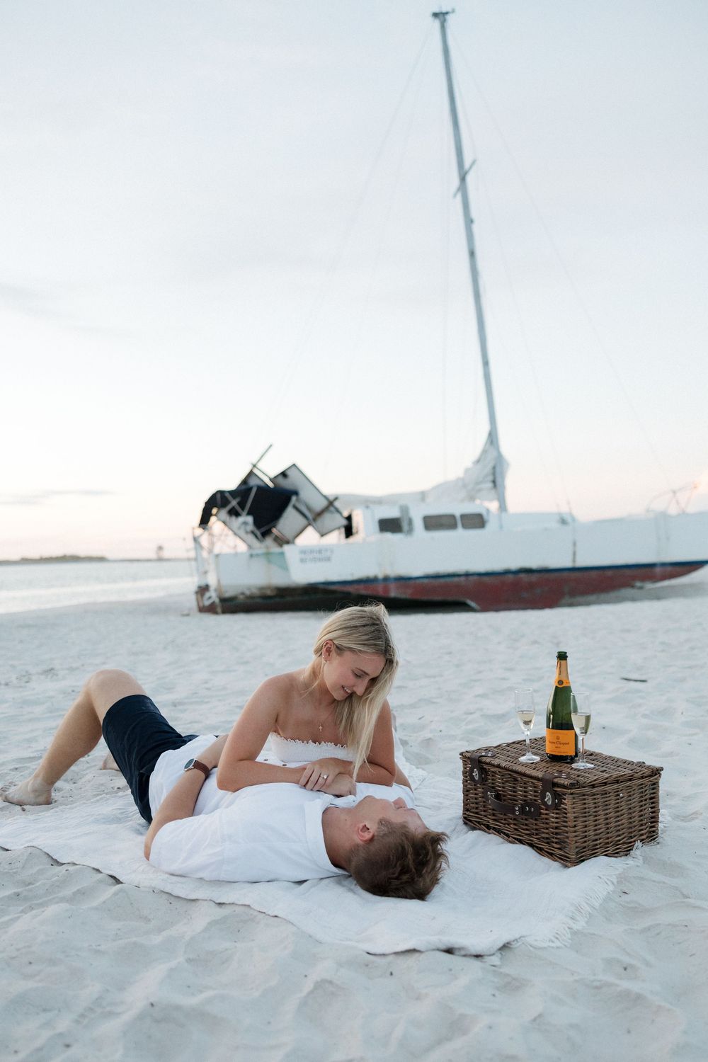 Couple relaxing on a beach blanket with a picnic basket and wine, with a sailboat in the background at sunset.