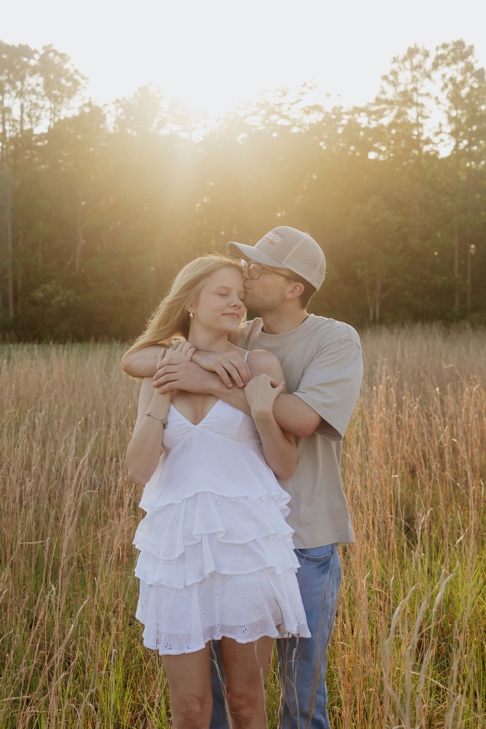 Couple embracing in golden field at sunset, man kissing woman in white ruffled dress.
