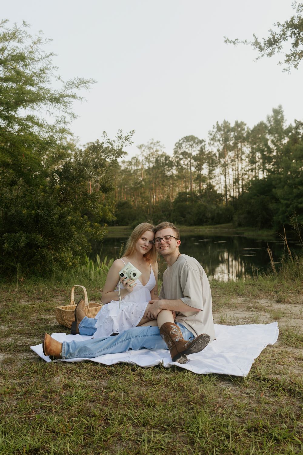 Couple sitting on white blanket for picnic by pond surrounded by tall pine trees.