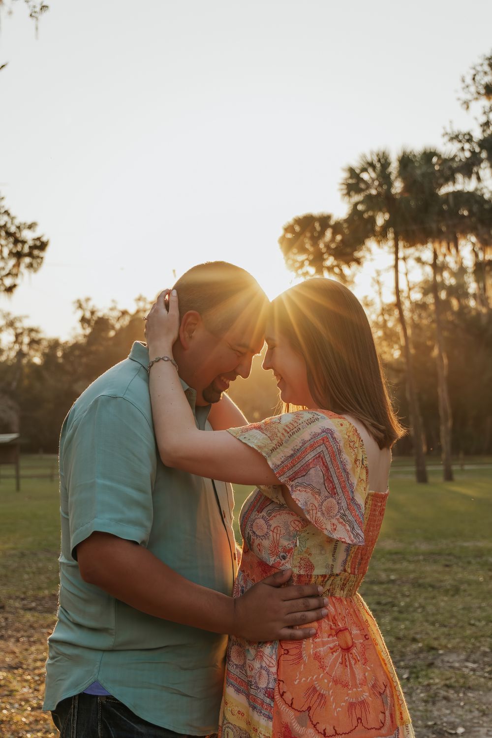 Couple sharing a romantic golden hour moment, foreheads touching, embraced in a sunlit park with palm trees.