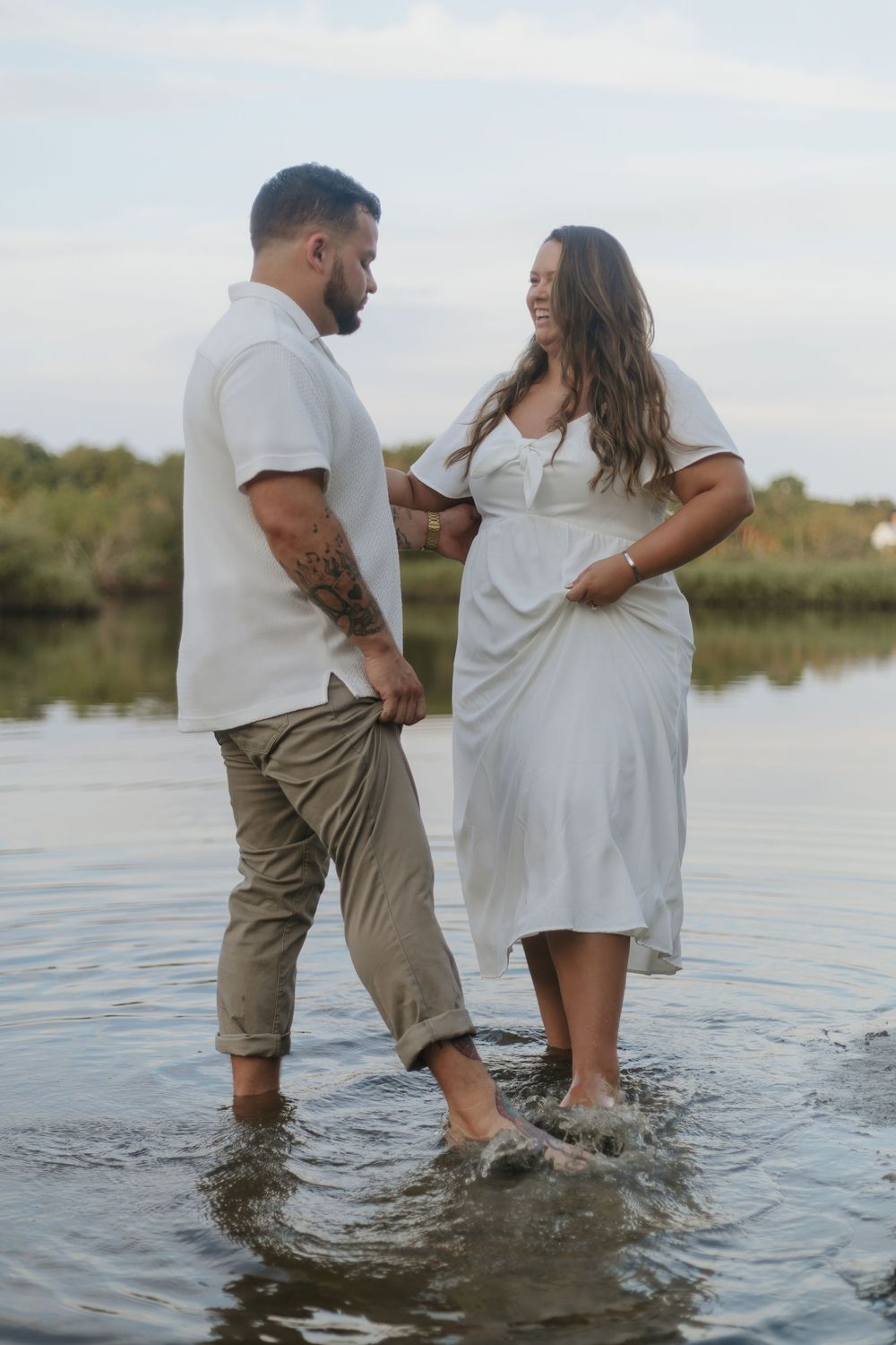 Couple wading in shallow water, woman in white dress laughing, man in white shirt and khaki pants, outdoor maternity photo.
