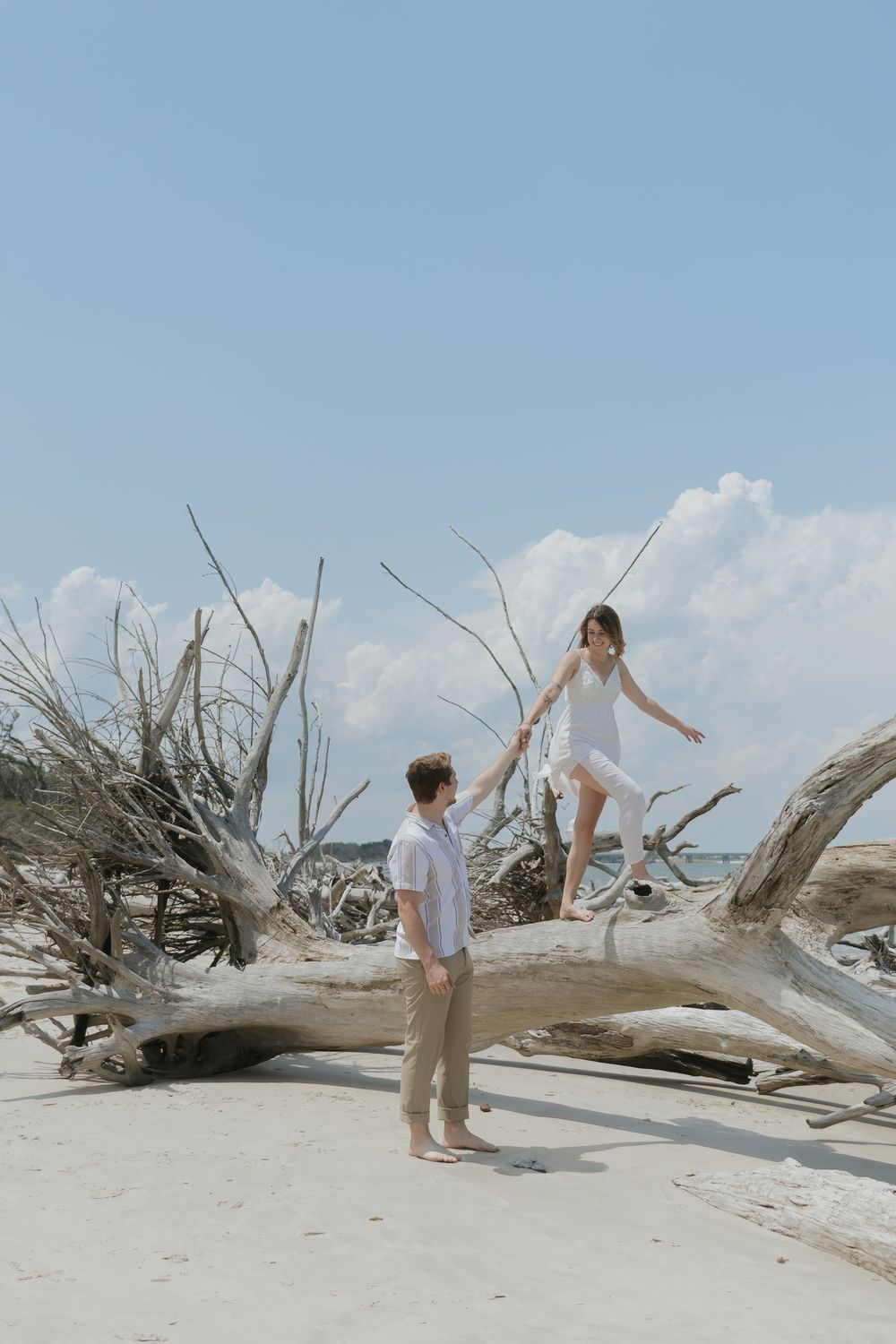 Couple on driftwood beach, woman in white dress balancing on log holding mans hand, blue sky backdrop.
