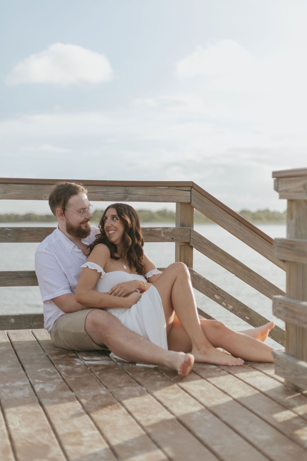 Happy couple sitting on wooden dock by water, woman in white dress laughing.