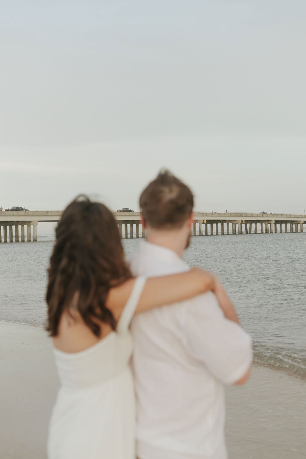 Couple in white embracing on beach, gazing at pier over calm water in soft focus romantic photo.