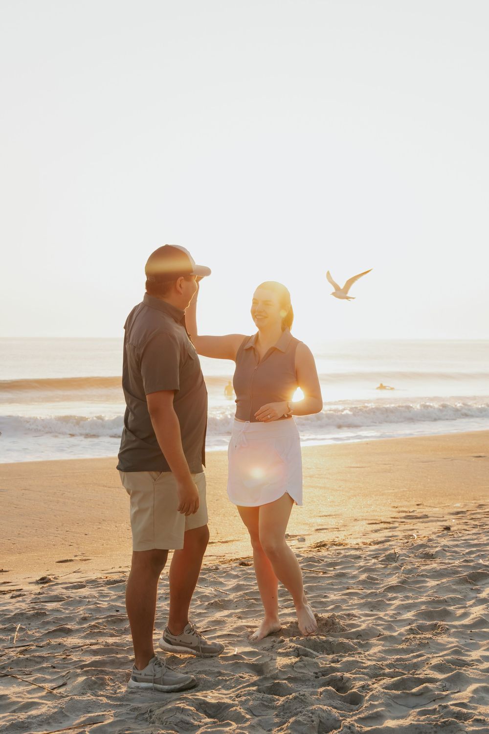 Couple standing on beach at golden sunset with seagull flying in background.