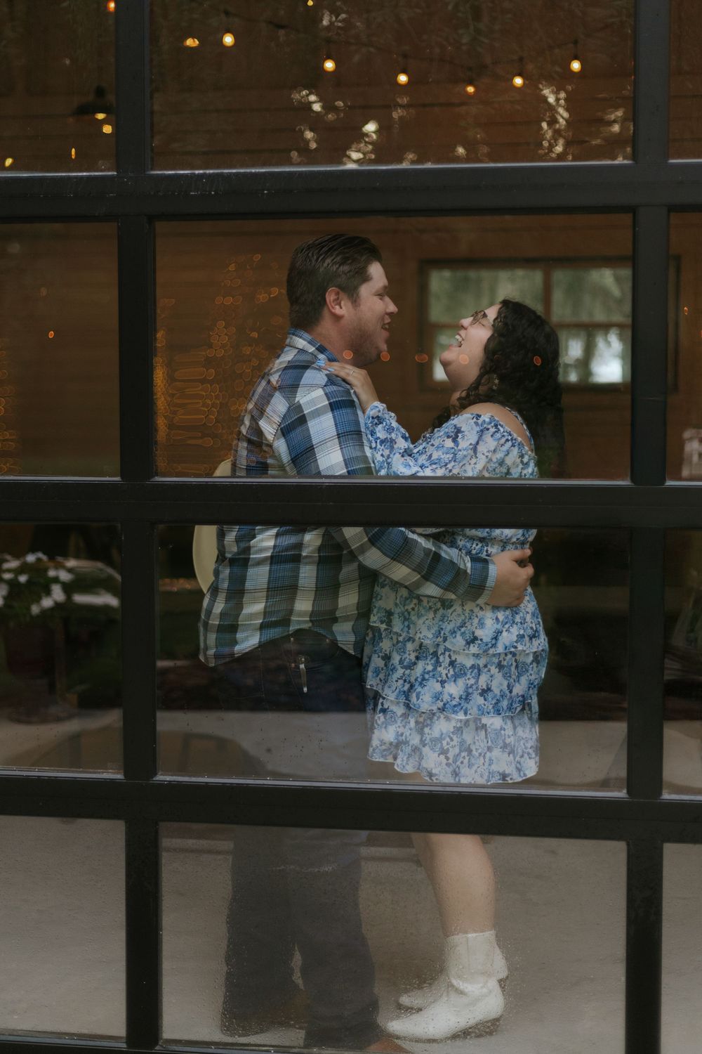 Couple embracing and laughing together through a glass window with warm string lights in background.