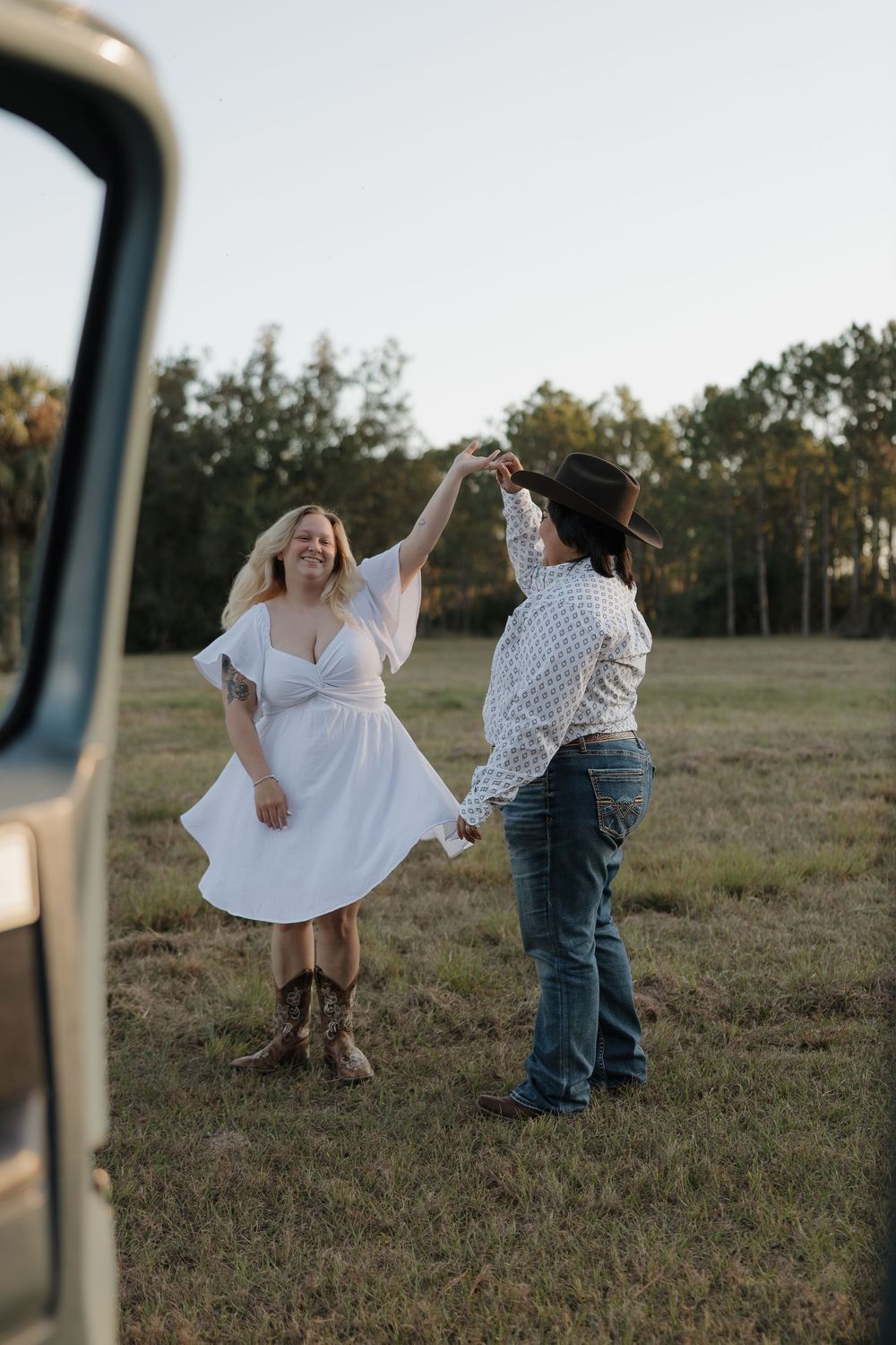 Couple dancing in an open field at sunset, woman in white dress and boots being twirled by partner in cowboy hat.