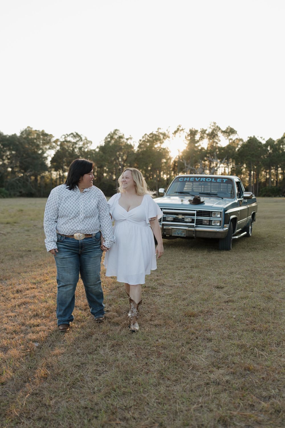 Two women stand together in a field at sunset beside a classic Chevrolet truck, one in jeans and one in a white dress.