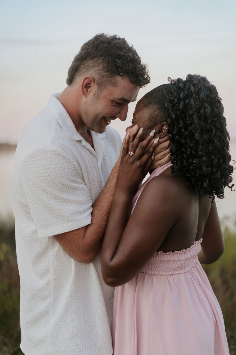 Interracial couple sharing a romantic moment outdoors, man in white shirt holding woman's face in pink dress at dusk.