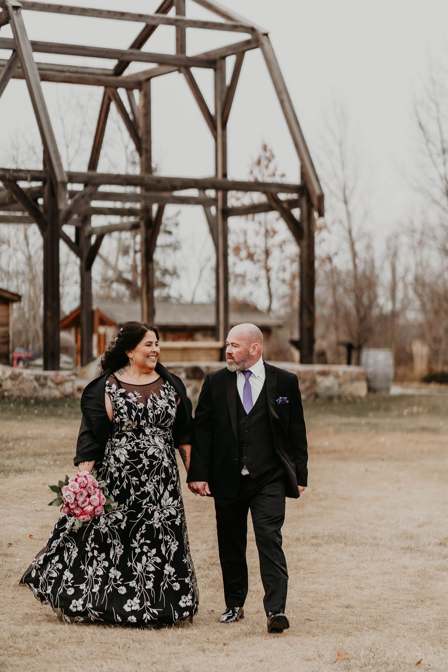 Couple walks hand-in-hand at rustic outdoor wedding venue, bride in black floral gown, groom in dark suit with lavender tie.