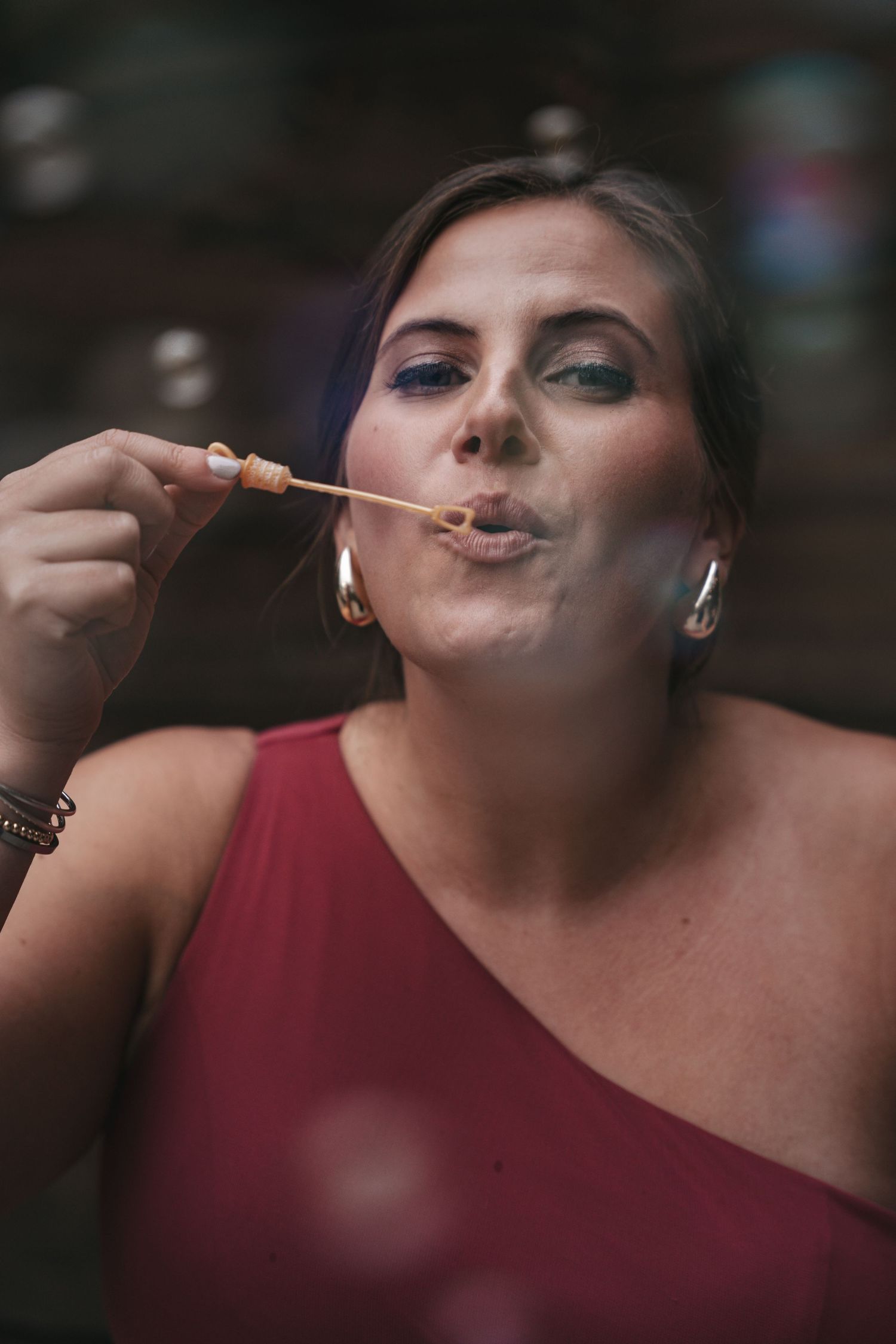 Woman in red dress blowing soap bubbles, wearing hoop earrings and colorful eye makeup.