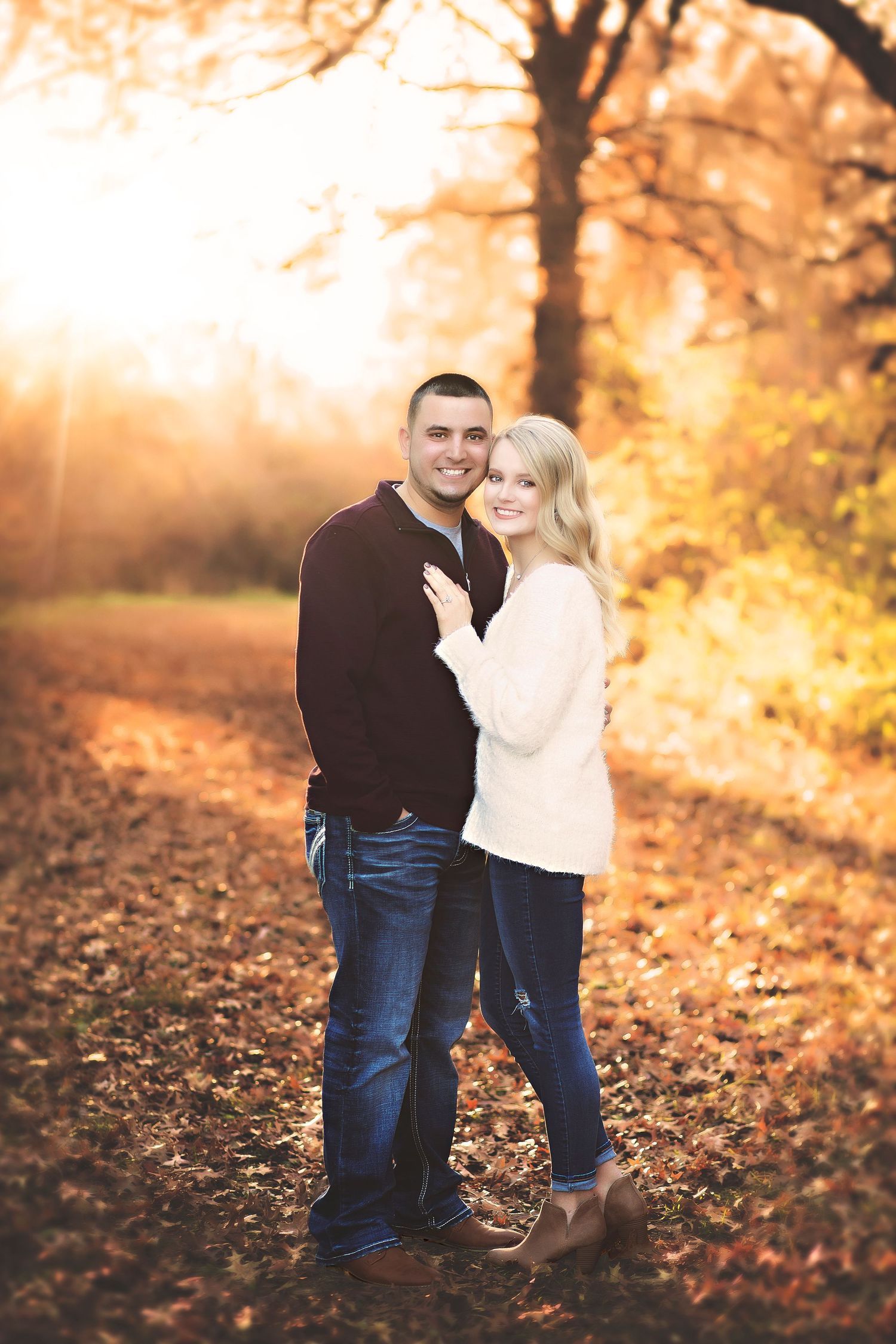 Couple embracing and smiling in a sunlit autumn forest, golden light filtering through bare trees onto fallen leaves.