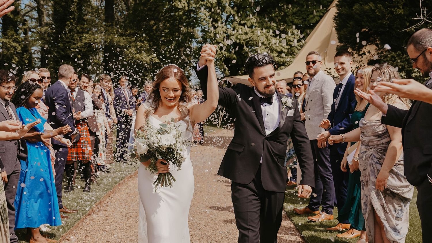 Bride and groom walk through confetti shower with guests cheering at outdoor wedding ceremony near tipi.