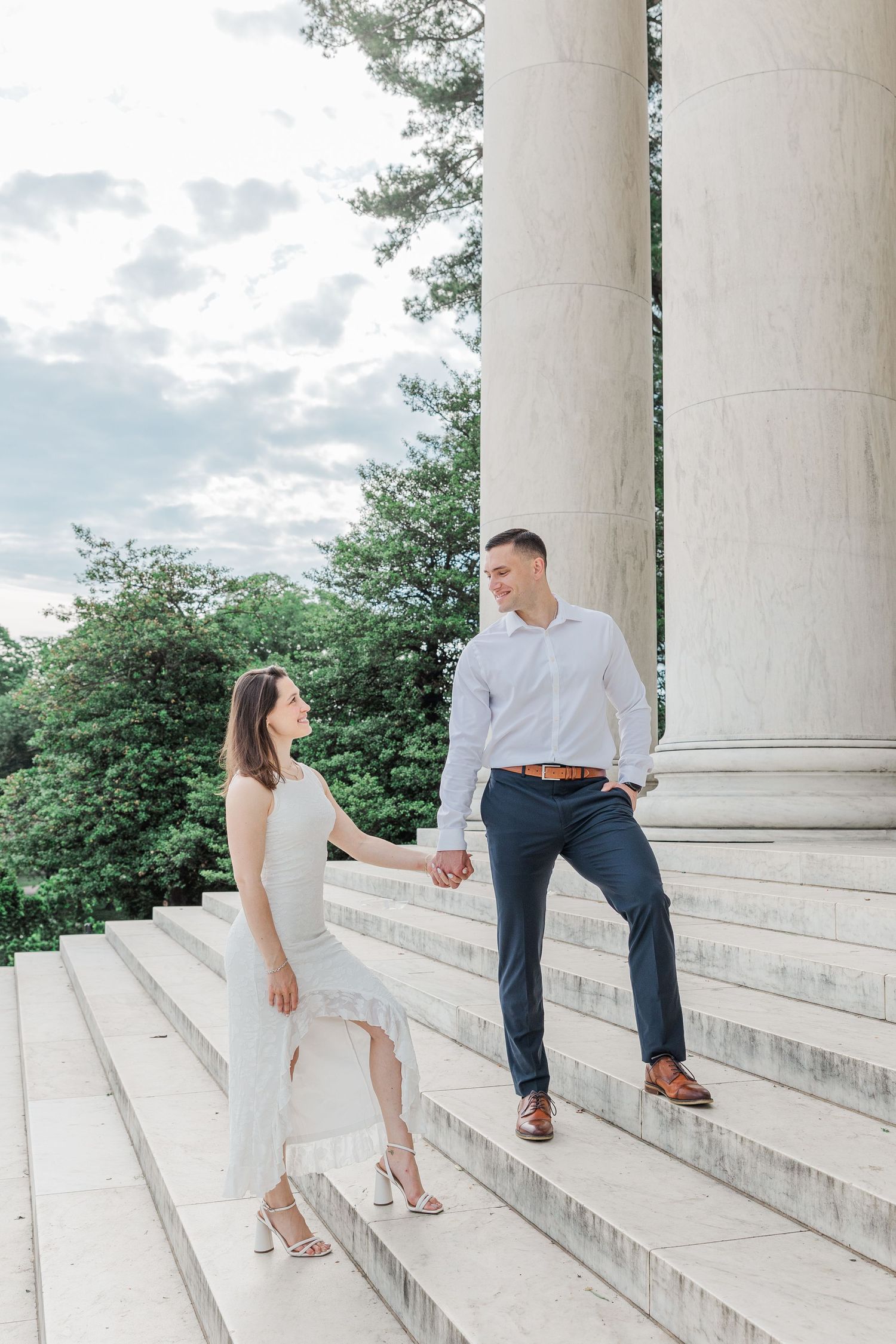 Couple holding hands on marble steps near large columns, woman in white dress and man in blue pants, outdoor engagement photo.