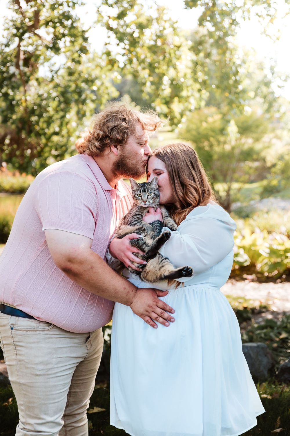 Couple kissing over a tabby cat they hold together outdoors in a sunny green garden setting.