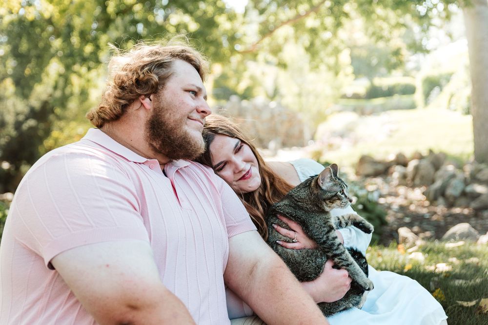 Couple sitting outdoors holding a tabby cat, smiling together in a sunny garden setting.