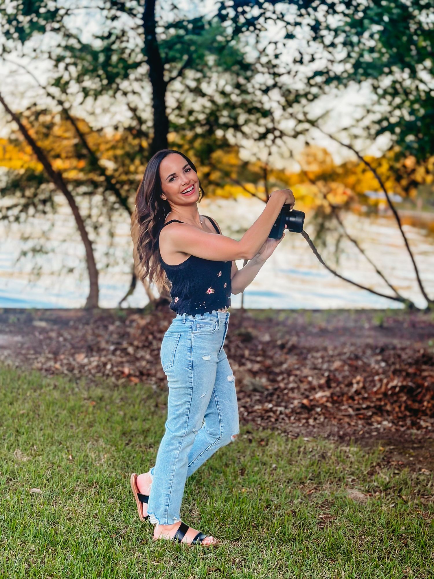 Smiling female photographer holding camera outdoors near trees during golden hour in casual jeans outfit.