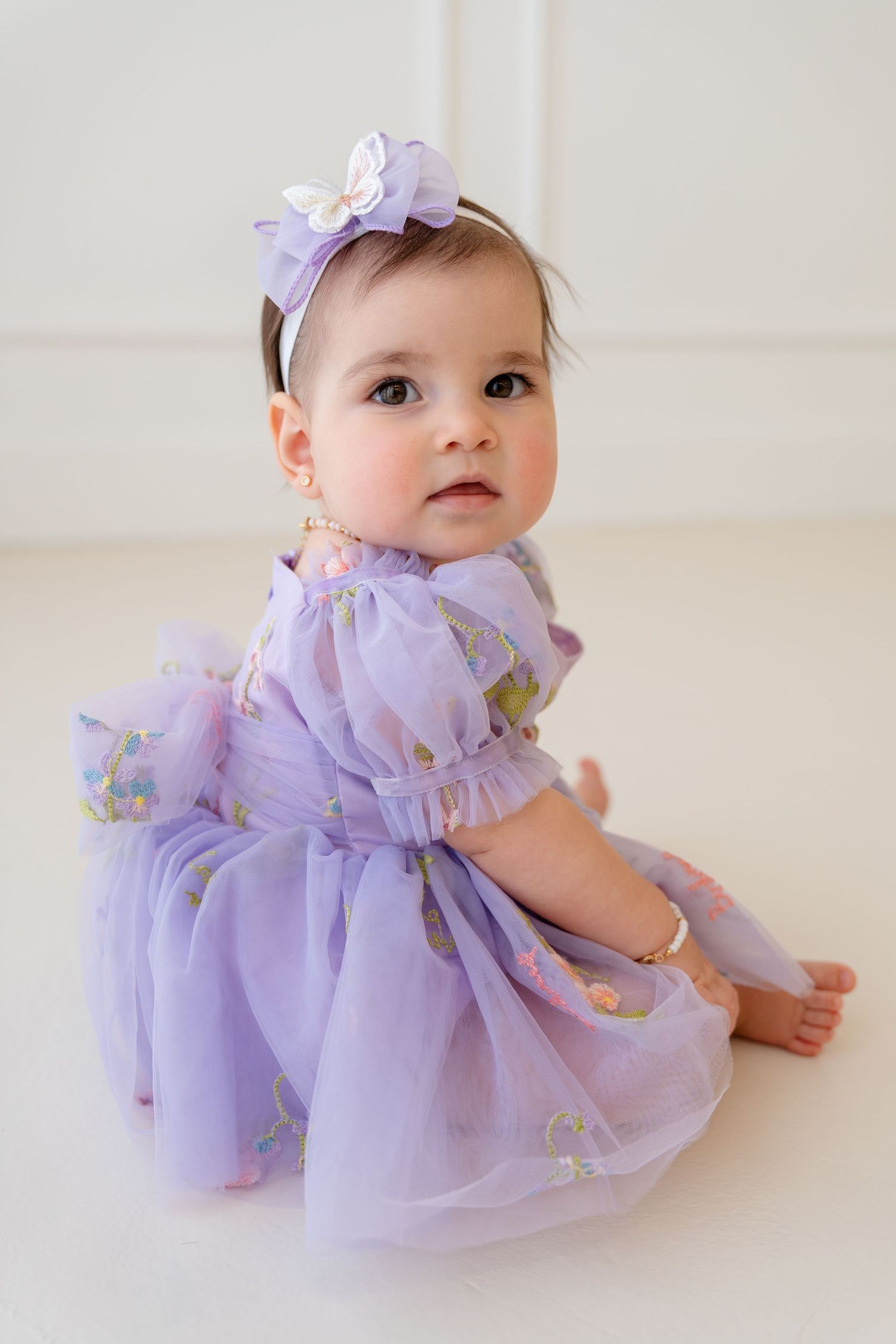 Baby girl in purple floral tulle dress with butterfly headband sitting on white floor.