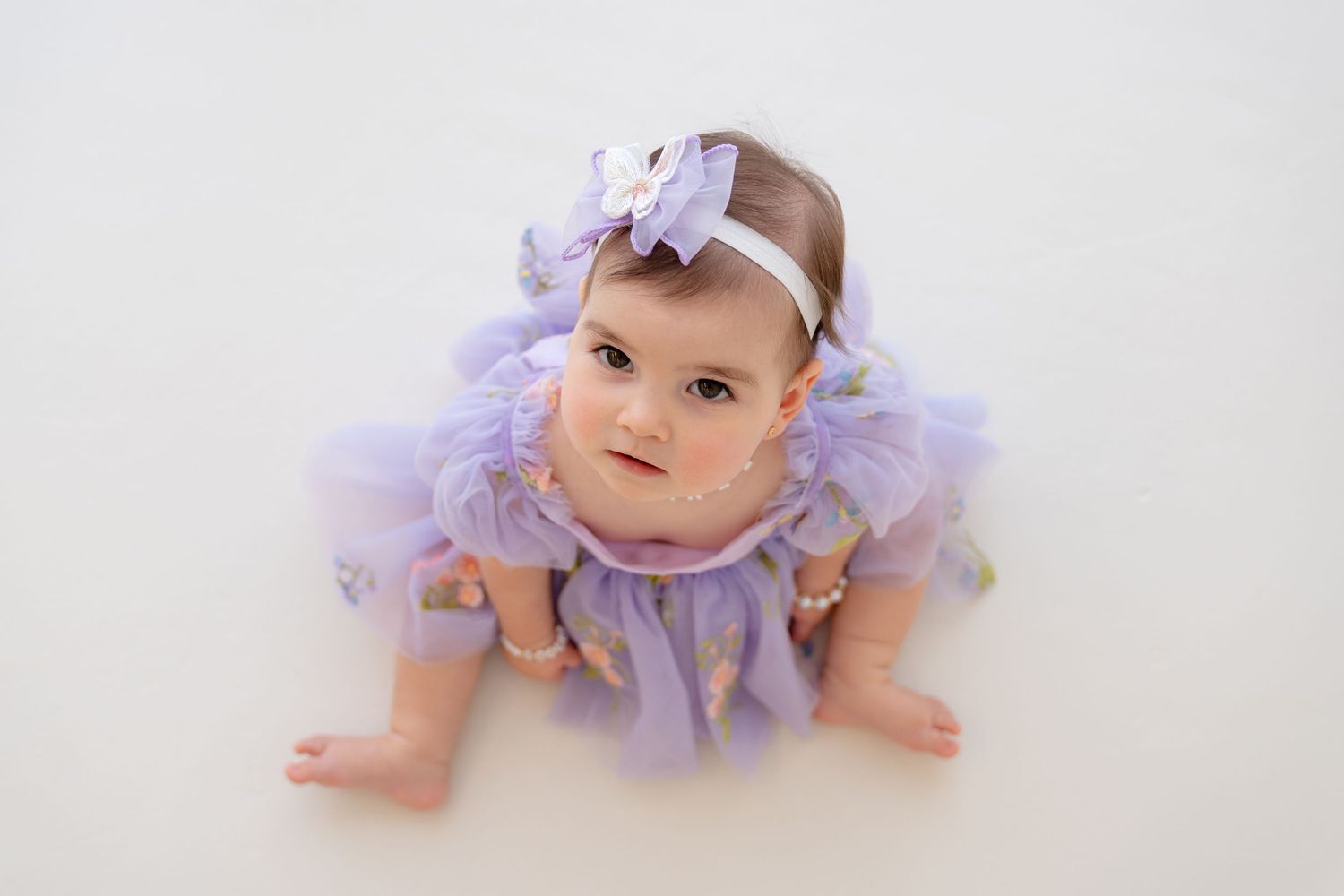 Adorable baby girl in lavender tutu dress and bow headband poses on white background.