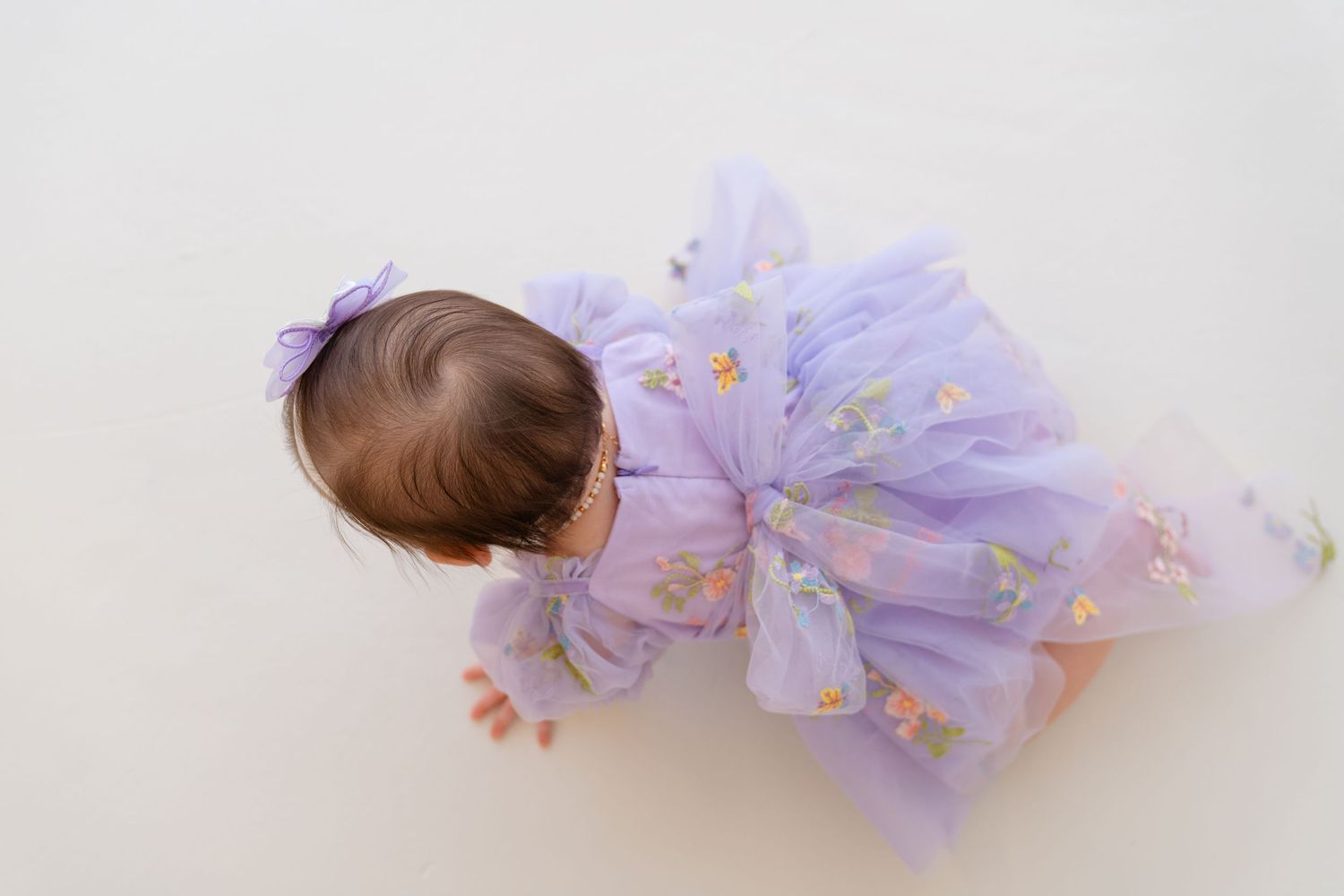 Baby girl in a lavender floral tulle dress with a purple bow, viewed from above on a white background.