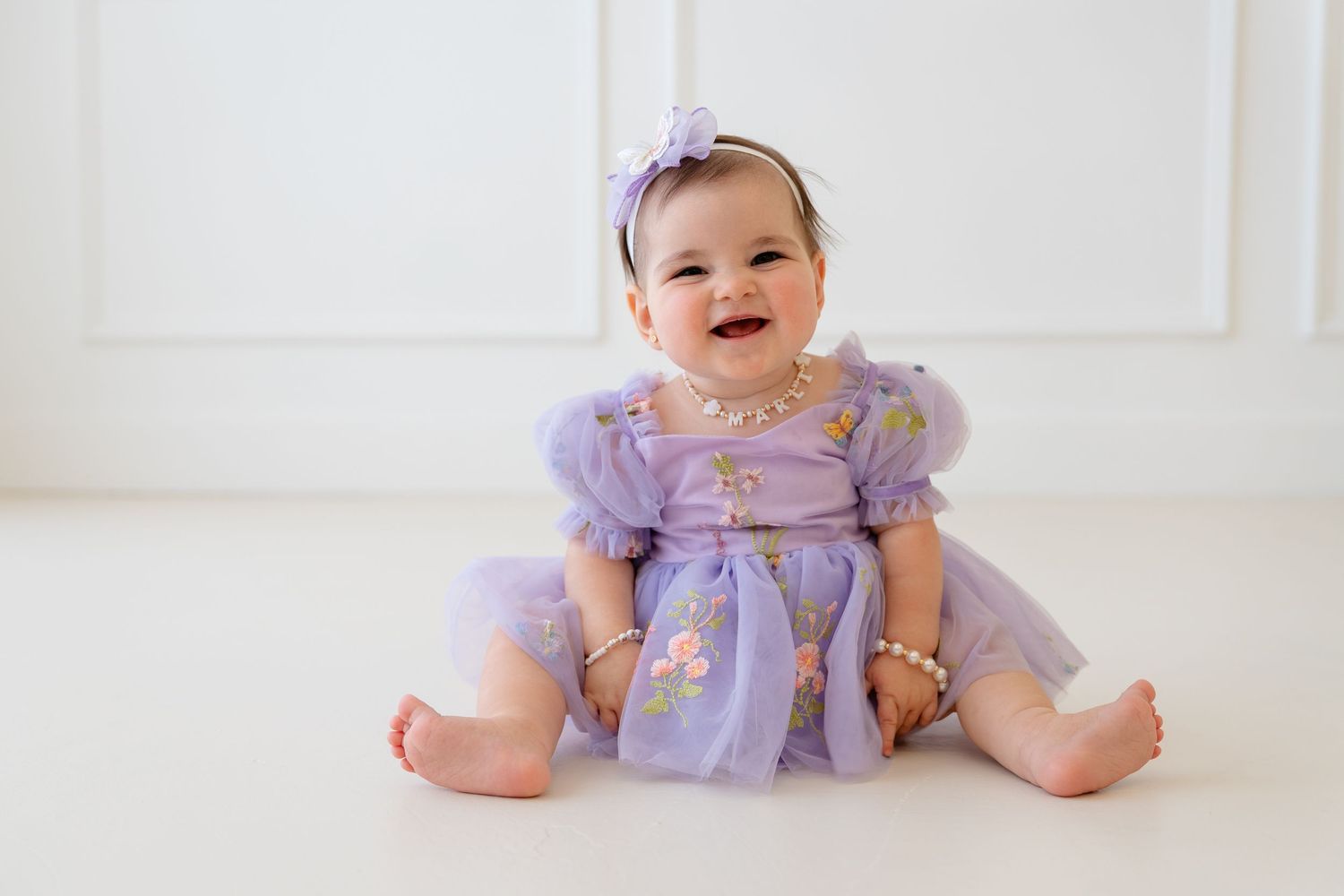 Smiling baby girl in lavender tulle dress with pearl necklace and floral headband sitting on white floor.