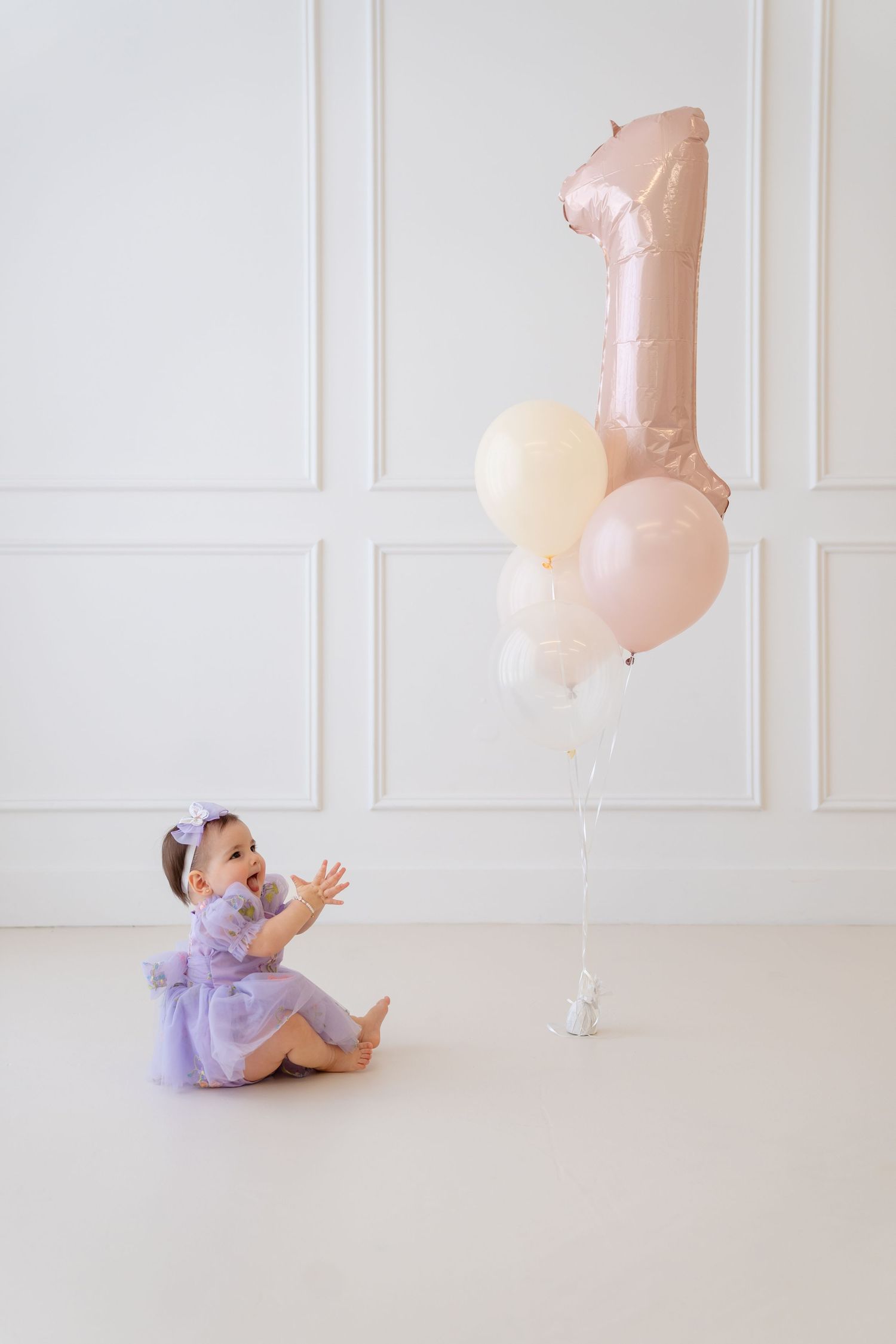 A baby girl in a purple dress sits on a white floor, looking up at colorful balloons and a number 1 balloon.