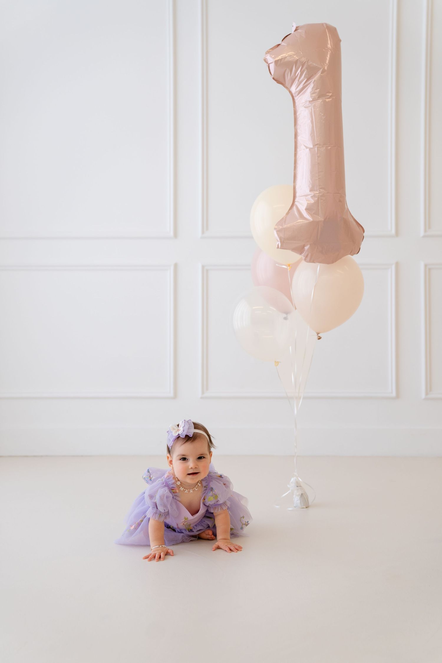 Baby girl in purple tulle dress crawls near number 1 balloon bouquet celebrating her first birthday.