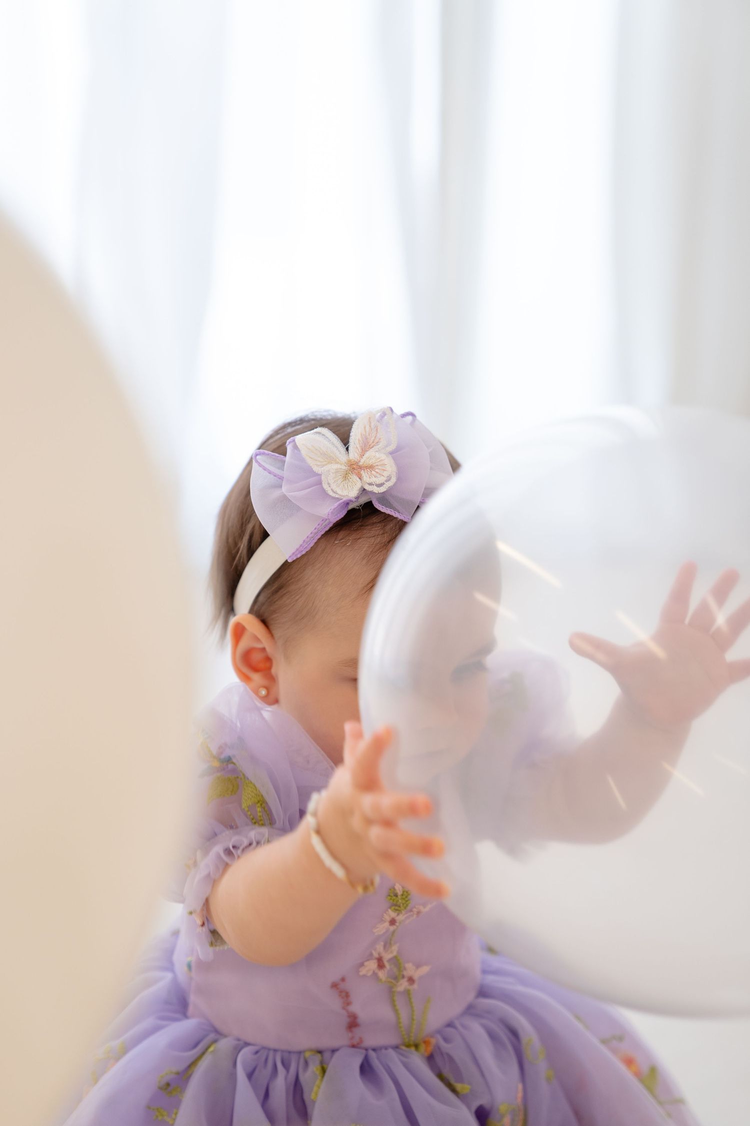 Baby girl in purple floral dress and bow headband playing with a white balloon near a bright window.