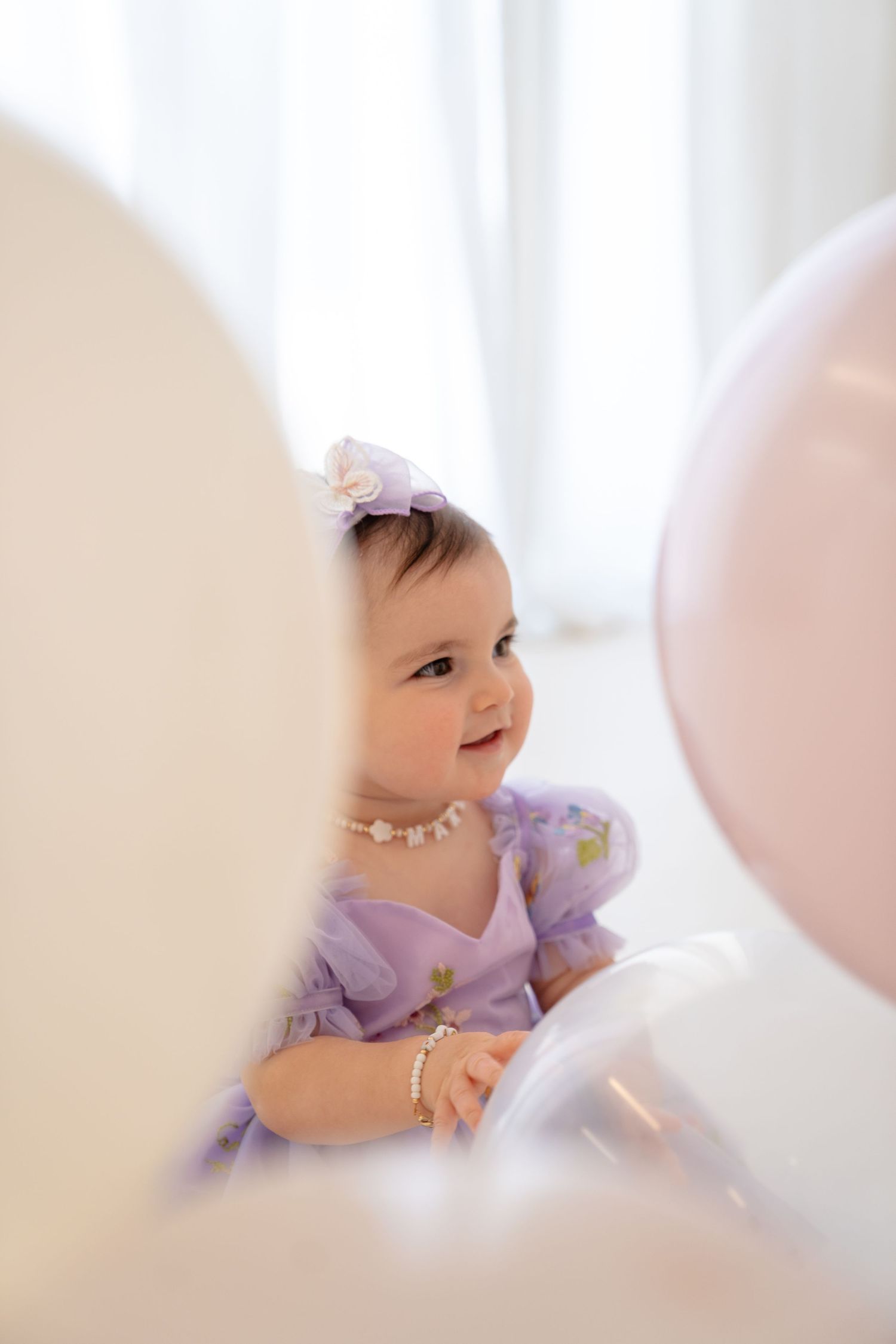 Baby girl in lavender dress with pearl necklace and bow headband surrounded by pink and white balloons.