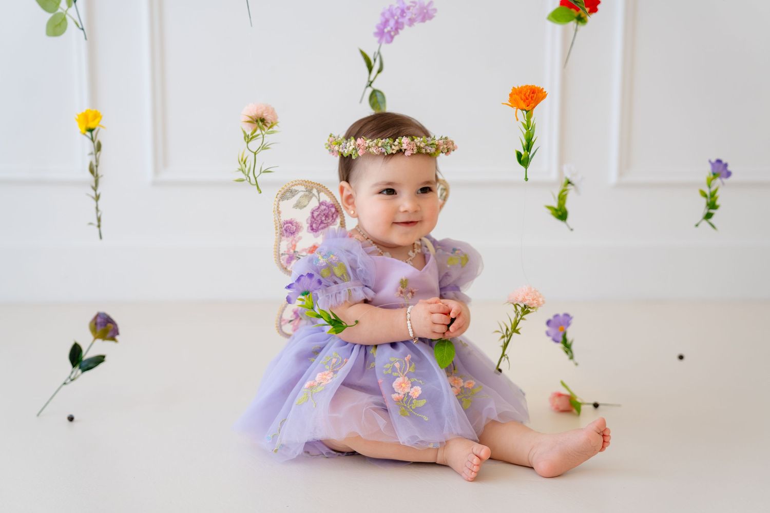 Baby girl in purple floral fairy dress and flower crown sits smiling among hanging flowers.