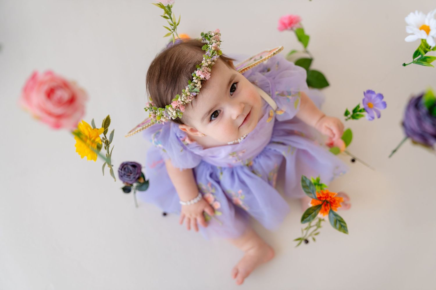 A baby girl in a purple tutu dress and floral crown sits surrounded by colorful flowers on a white background.