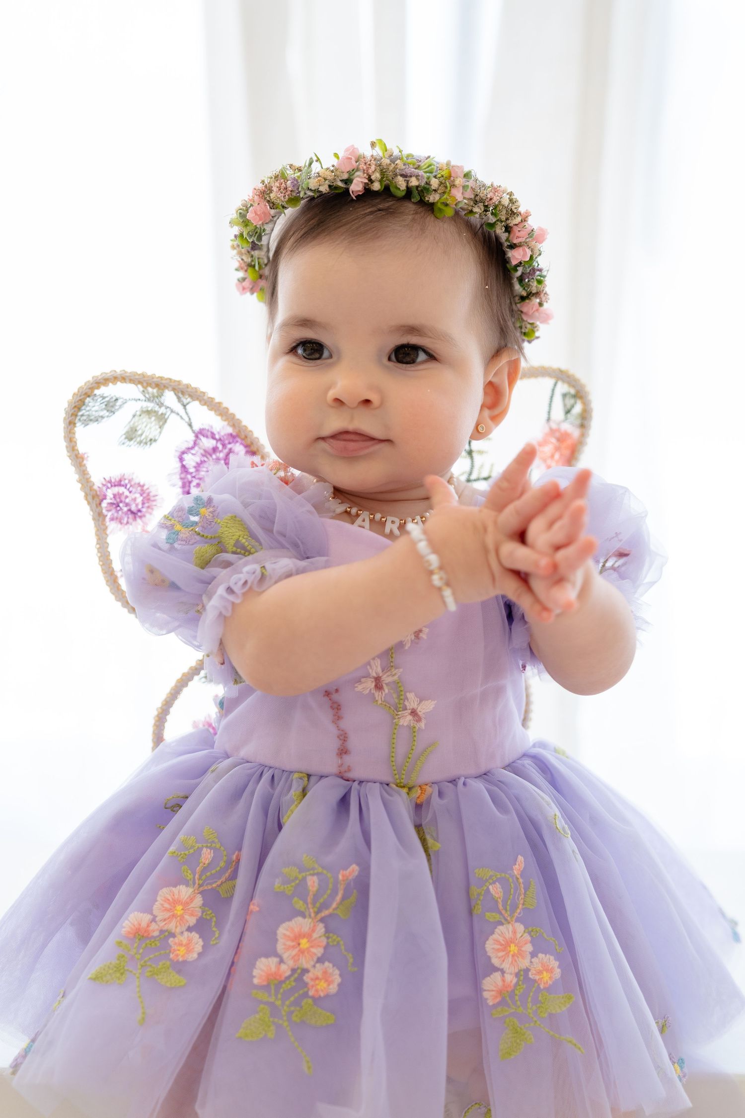 Baby girl in purple floral dress with fairy wings and flower crown reaches forward.