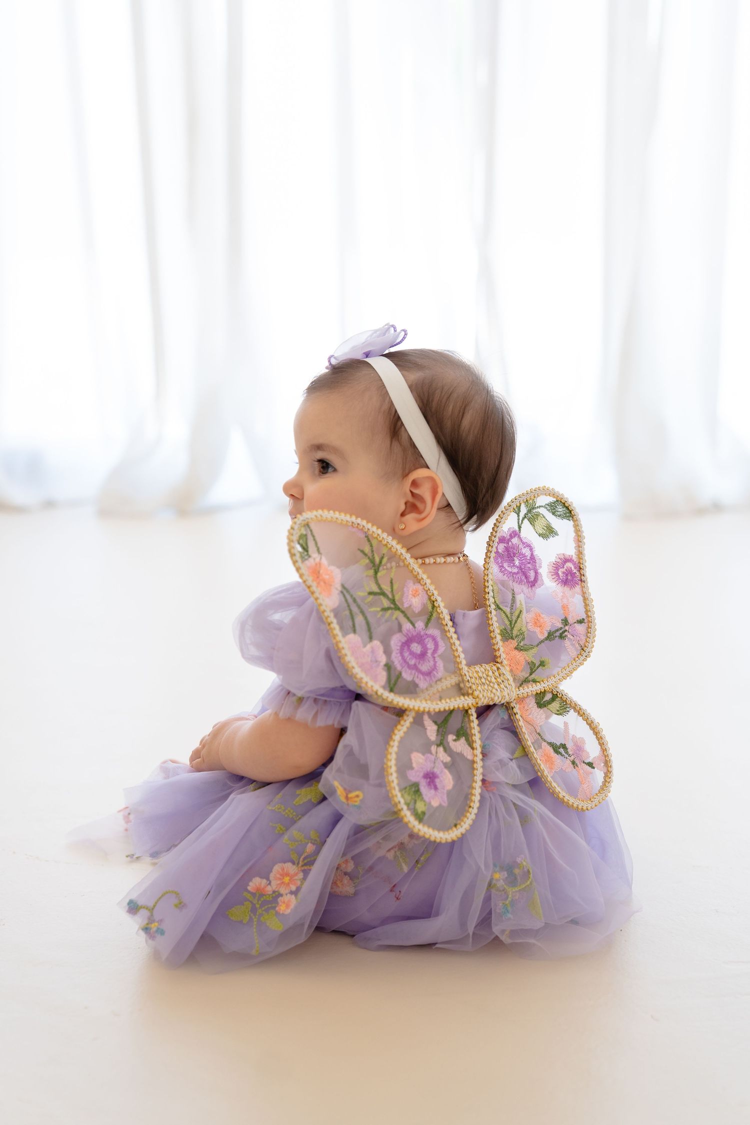 Baby girl in purple tulle dress with floral butterfly wings sits on white floor for first birthday.