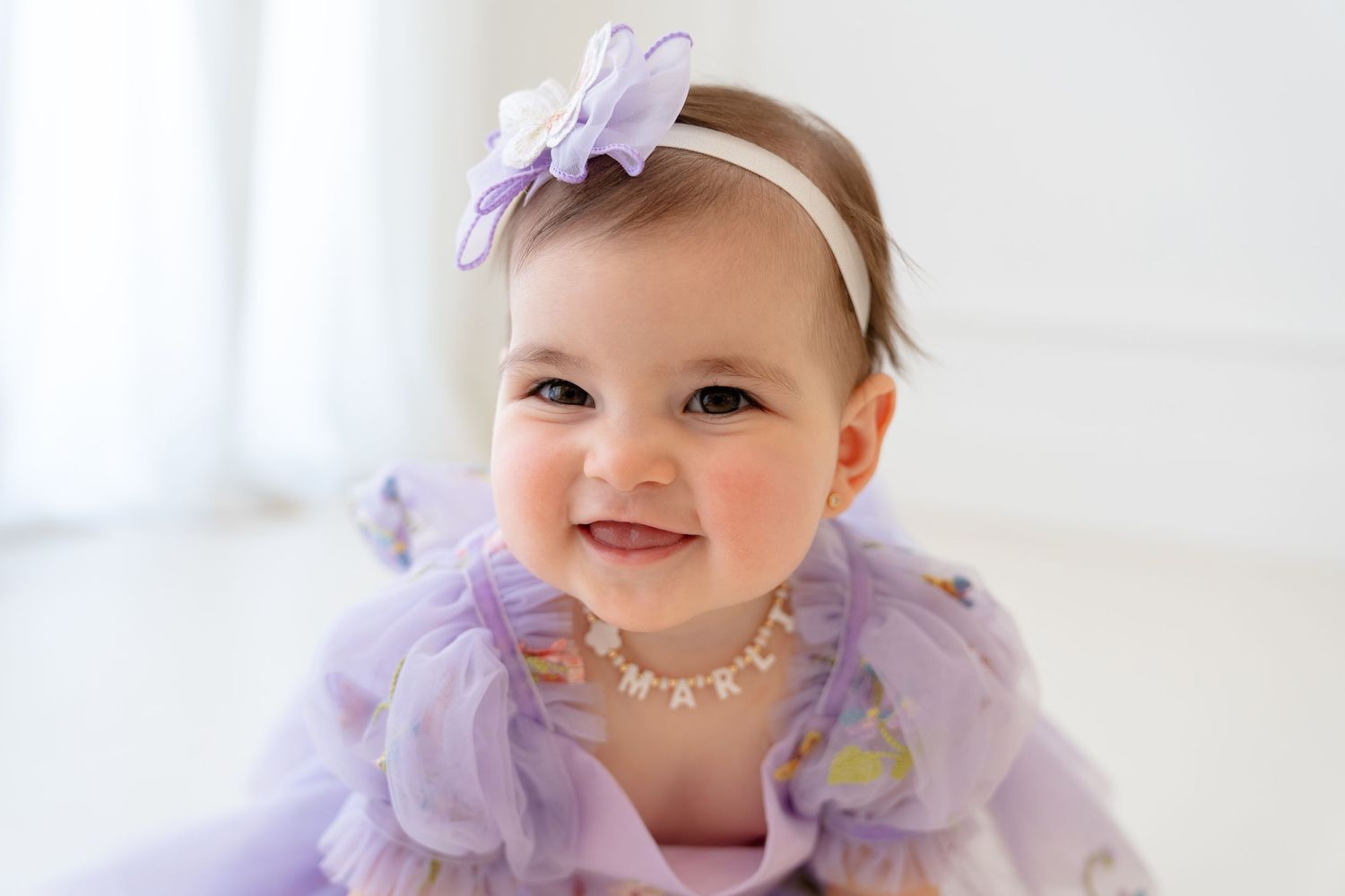 Smiling baby girl in purple tutu dress and bow headband wearing a name necklace, bright white studio photo.