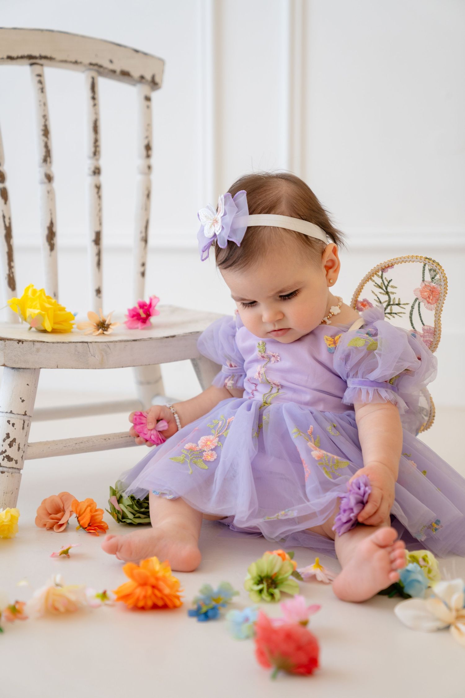 Baby girl in purple fairy dress sits on floor surrounded by colorful flowers near white chair.