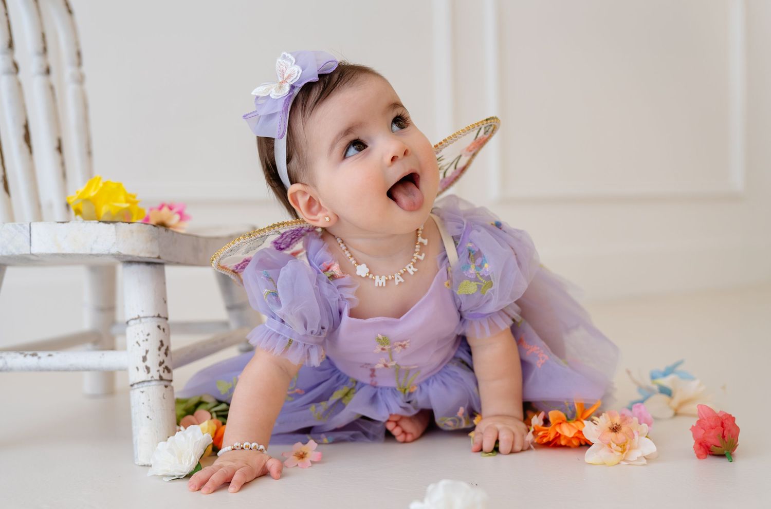 Baby girl in purple tutu dress and pearl necklace sits on floor surrounded by flowers, mouth open in delight.