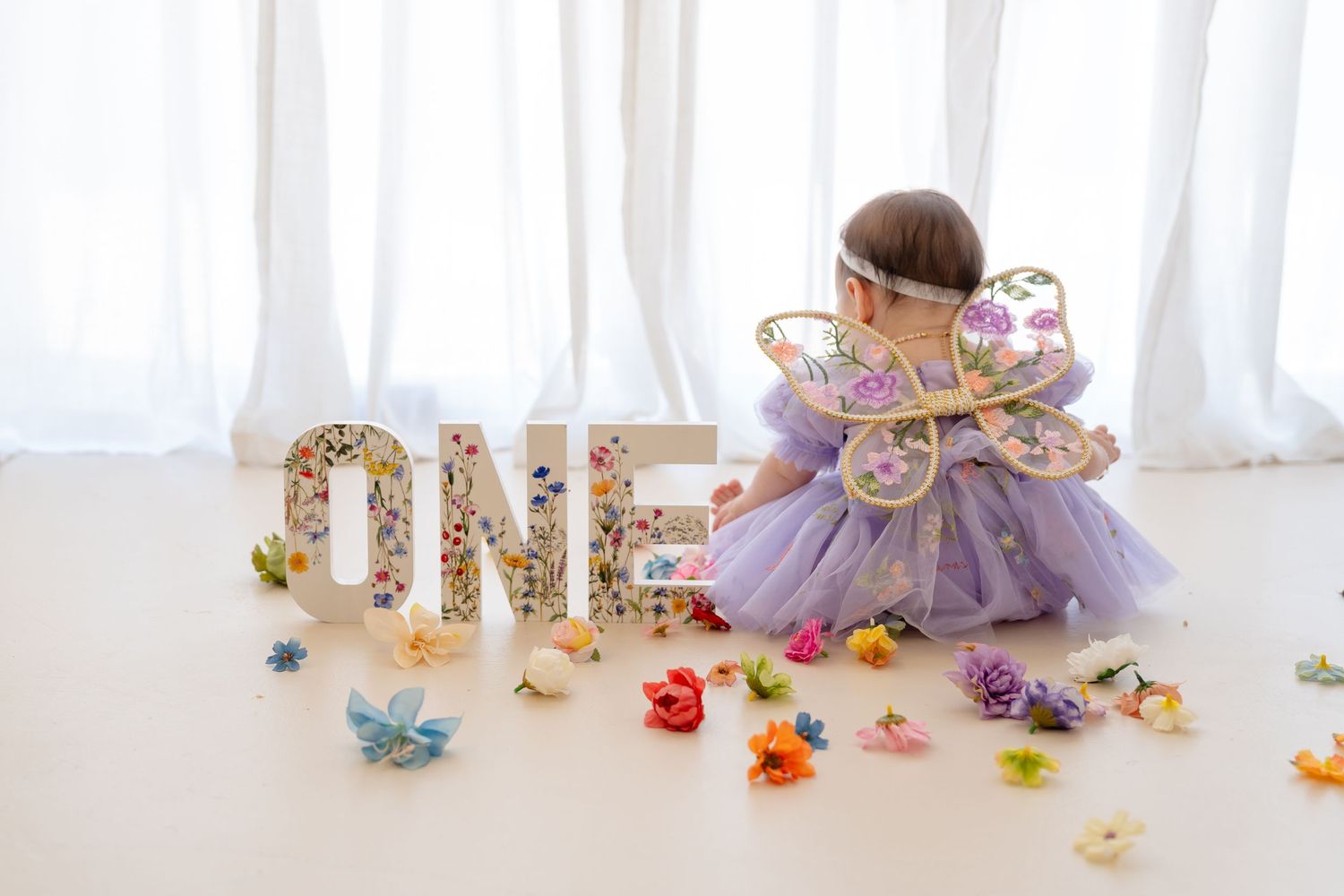 Baby girl in purple fairy dress with butterfly wings sits beside floral ONE sign surrounded by colorful flowers.