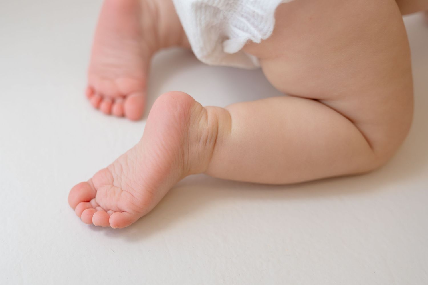 Close-up of chubby baby feet and legs resting on a white surface, wearing a white diaper.