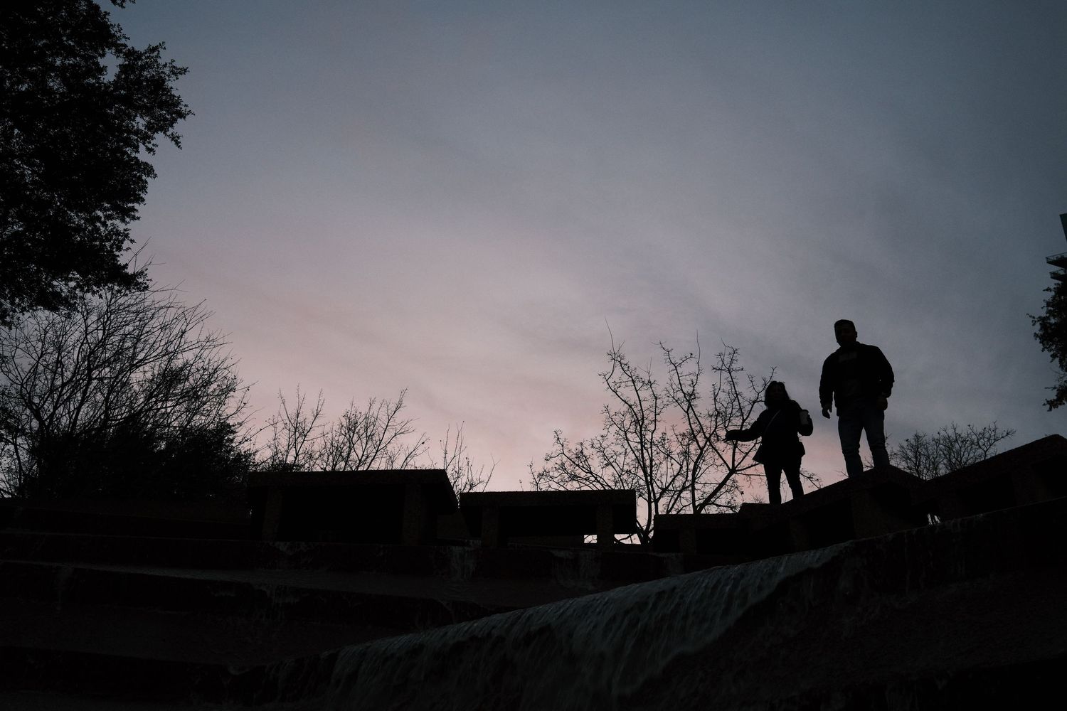 Silhouettes of two people standing on a wall at dusk with bare trees and a colorful twilight sky.