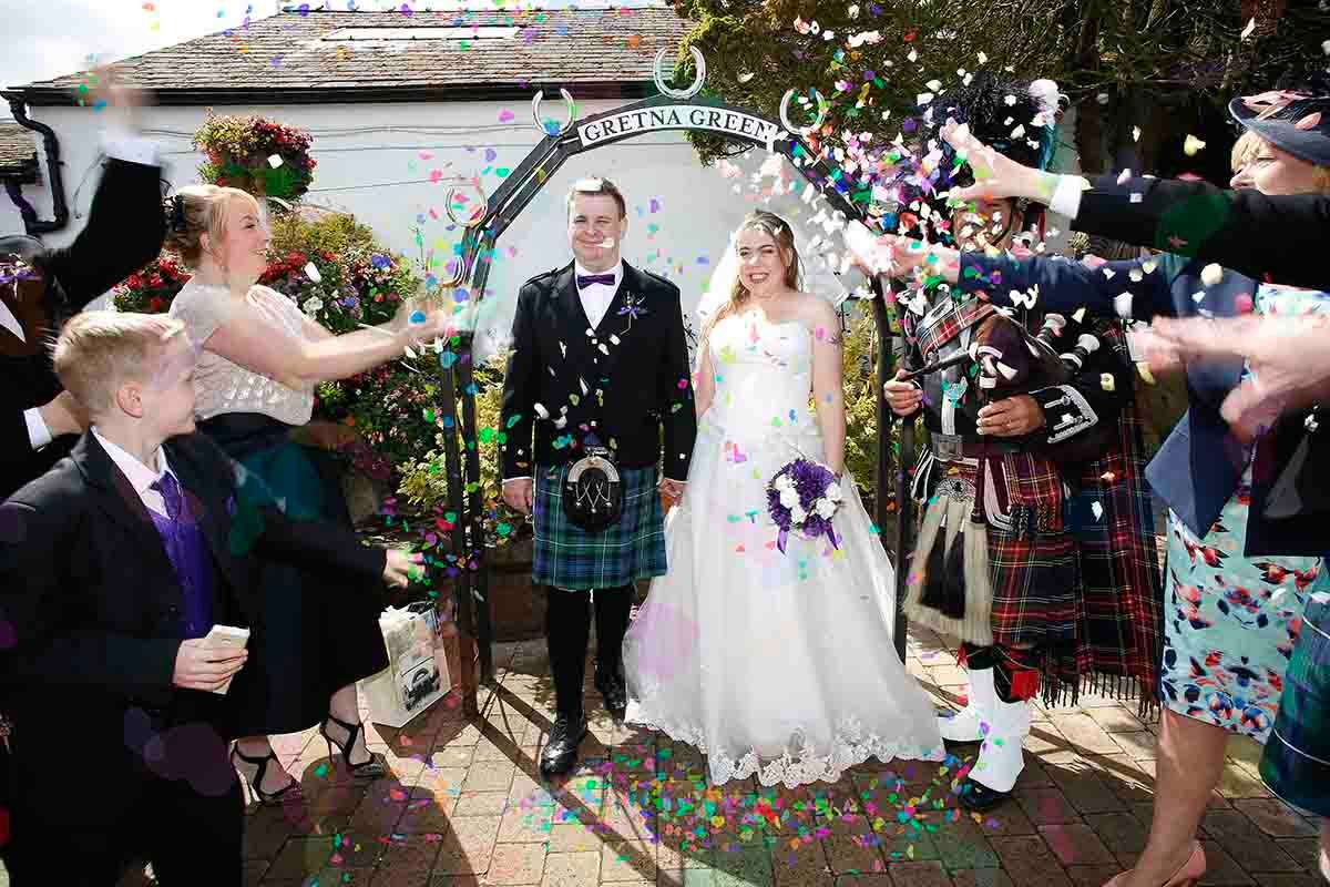 Scottish wedding couple in kilts and white gown celebrate with colorful confetti thrown by guests outside The Old Smithies in Gretna