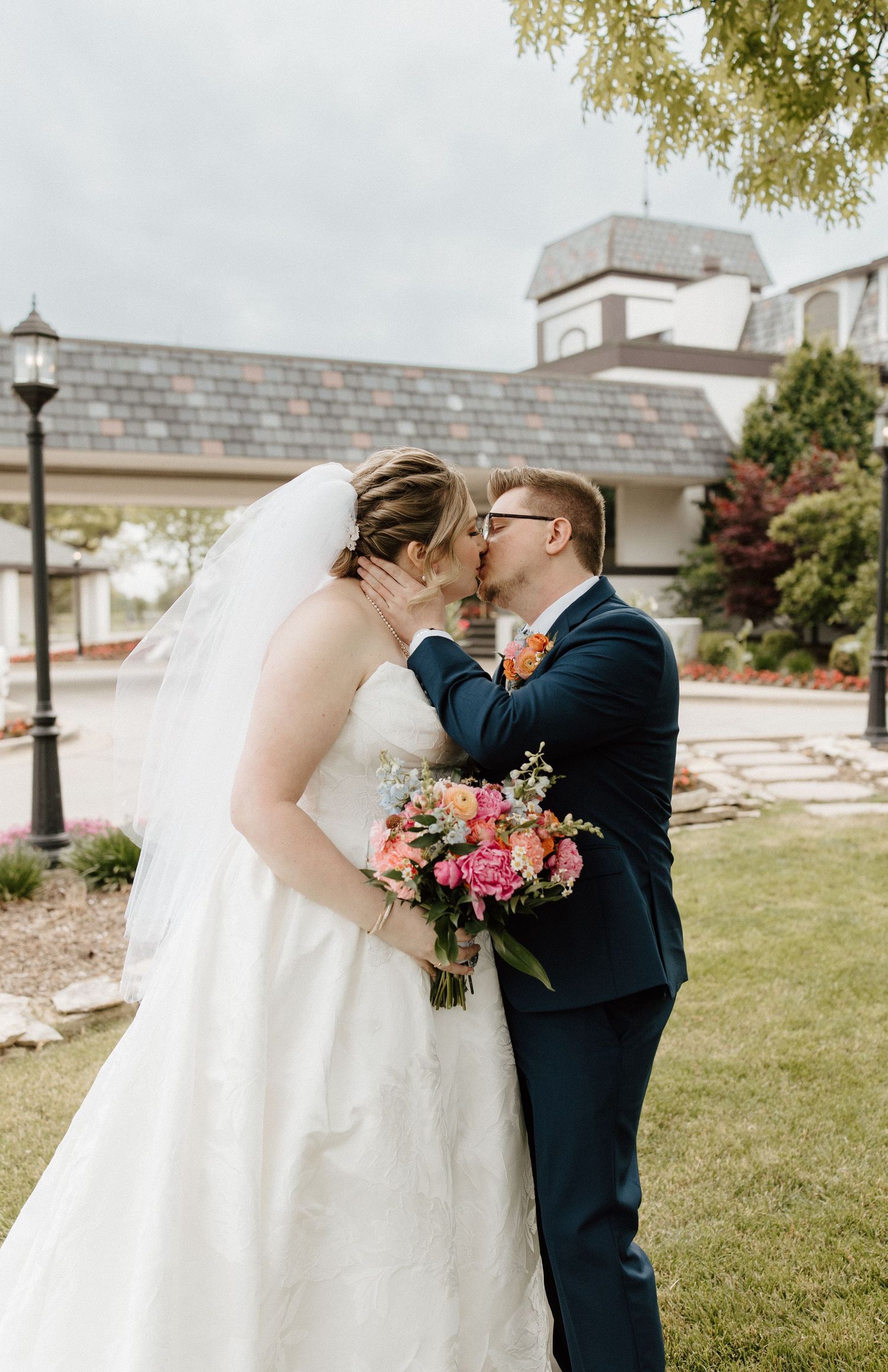 A bride and groom share a tender forehead touch outdoors, dressed in a lace gown and gray suit, surrounded by bare trees.