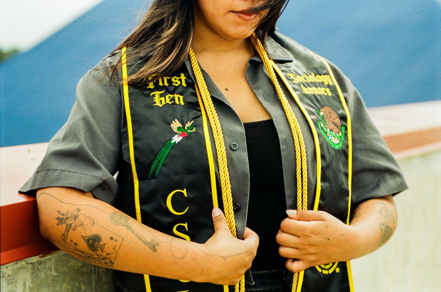 Graduate wearing black and gold First Gen stole with tattoos visible, posing outdoors in graduation attire.