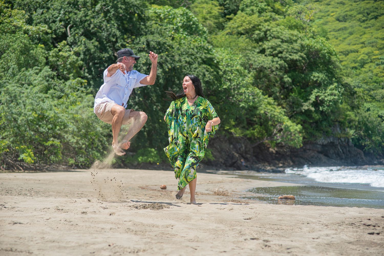Elderly couple joyfully playing on a tropical sandy beach, man jumping in the air, woman in green floral outfit.