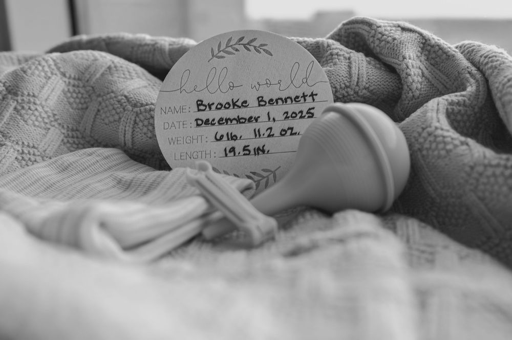 Black and white newborn photo with birth announcement sign for Brooke Bennett, born December 1, 2025, 6lbs 11.2oz, 19.5in.