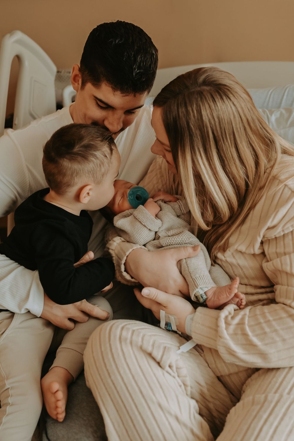 A young family tenderly gazes at their newborn baby in a hospital room, with toddler sibling leaning in to kiss.
