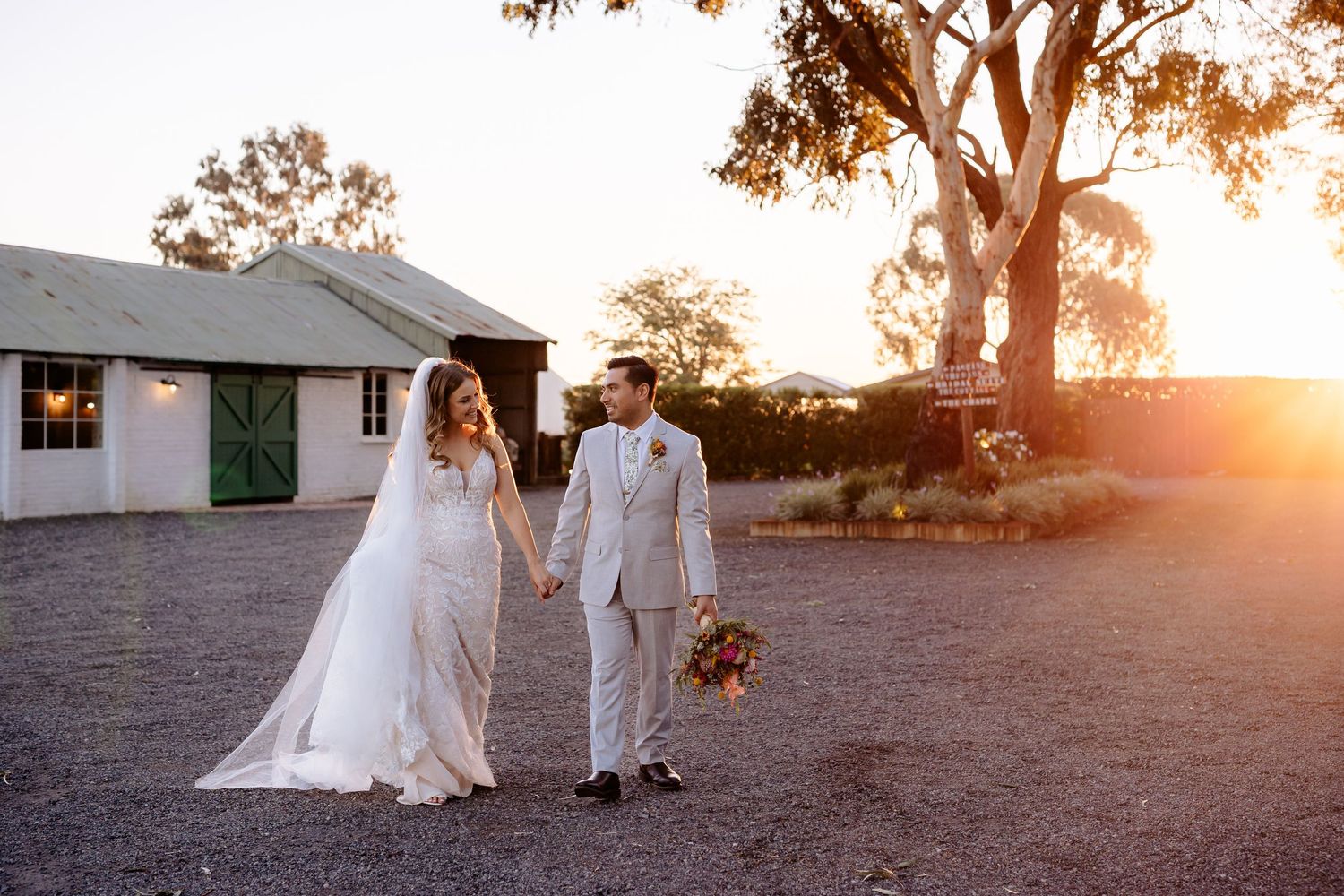 Bride and groom walk hand-in-hand at golden sunset near rustic barn venue on their wedding day.