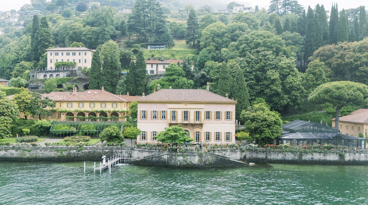 Elegant Italian villa on Lake Como shoreline surrounded by lush green trees and hillside gardens.
