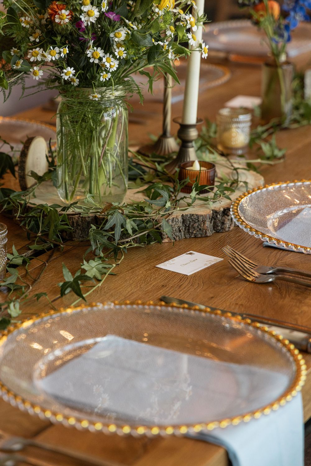 Rustic wedding table setting with gold-rimmed charger plates, wildflower centerpiece in mason jar, candles, and greenery on wood table.
