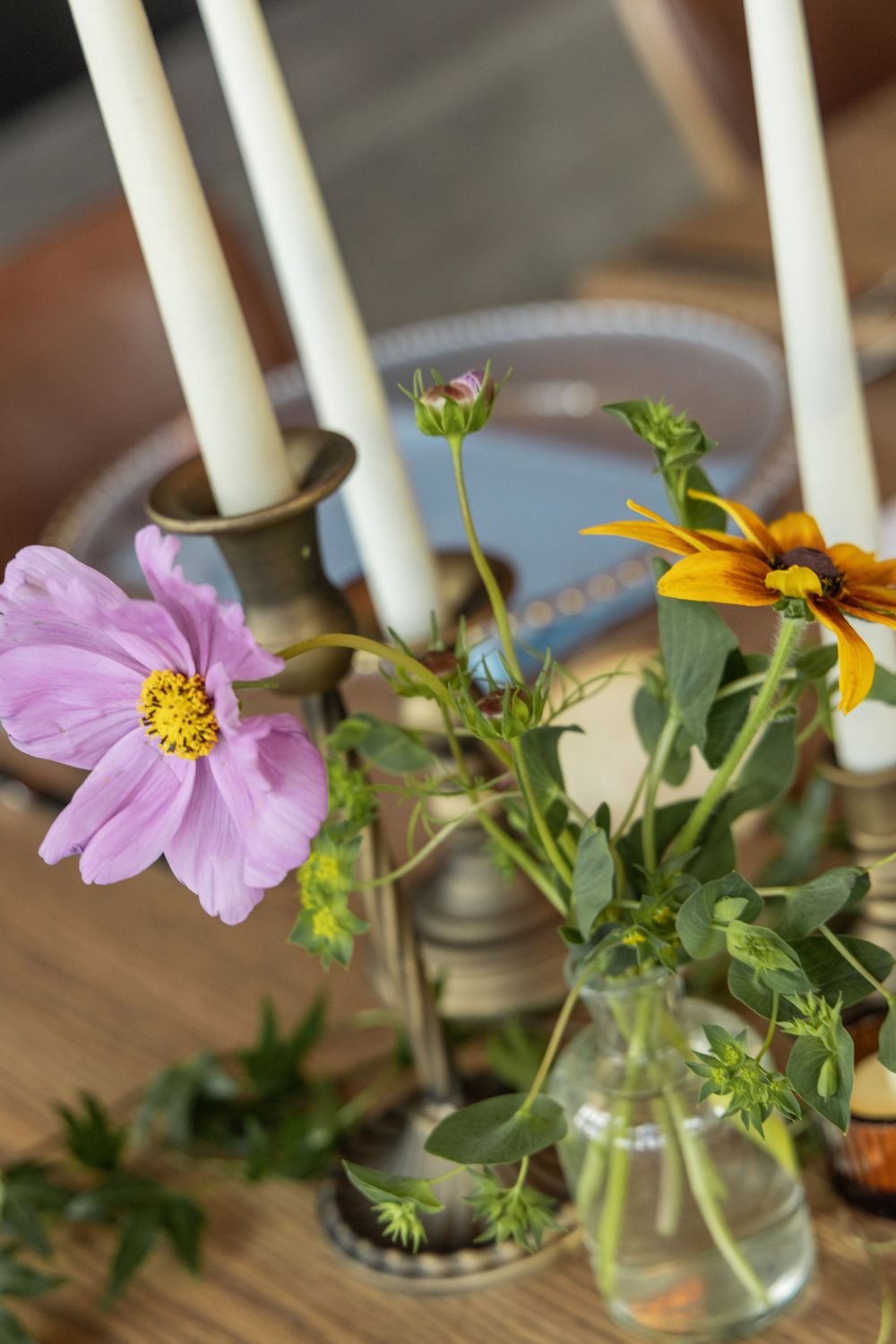 Vintage brass candelabra with white candles beside pink cosmos and yellow rudbeckia flowers in glass vases on wooden table.